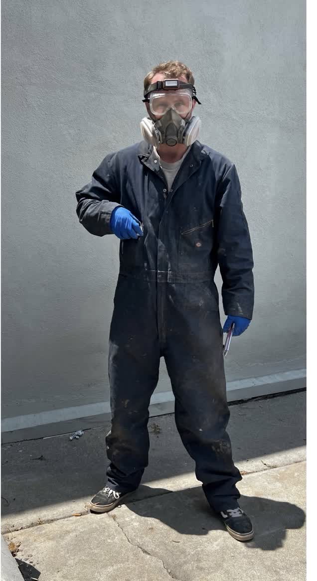 Plumbing technician in coveralls wearing a respirator, safety goggles, and protective gloves