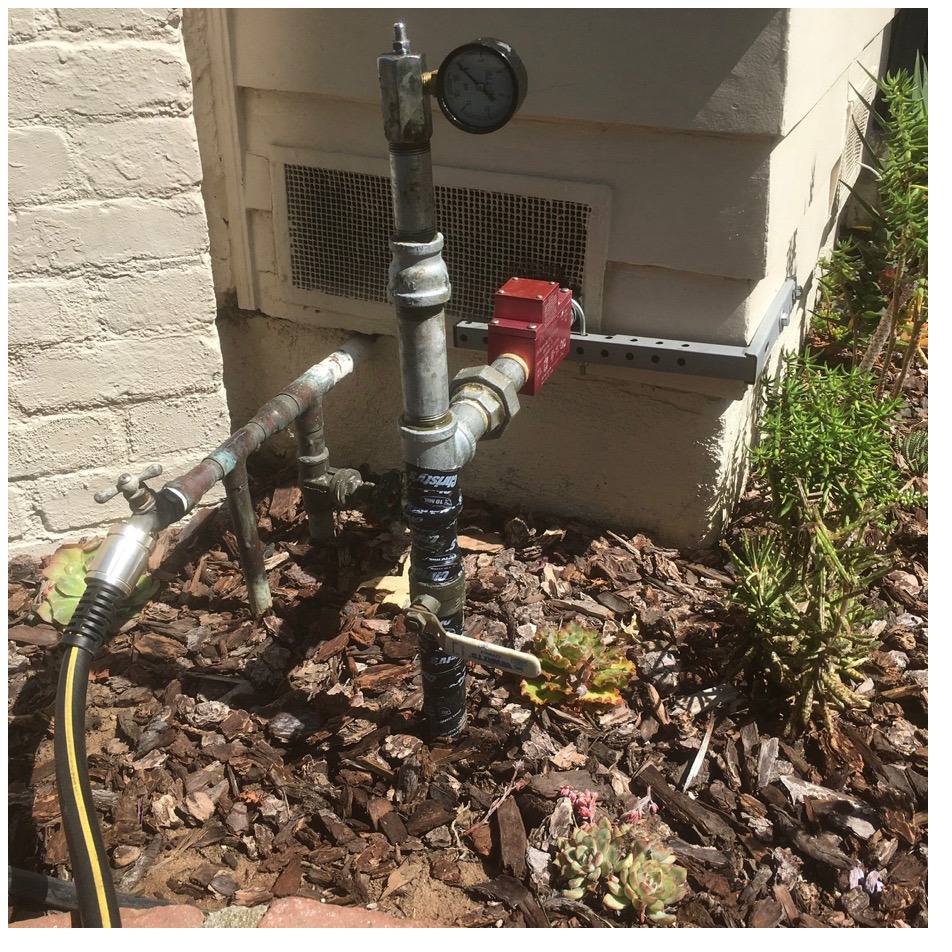 Red earthquake safety valve on exterior gas piping with a pressure gauge beside a brick wall