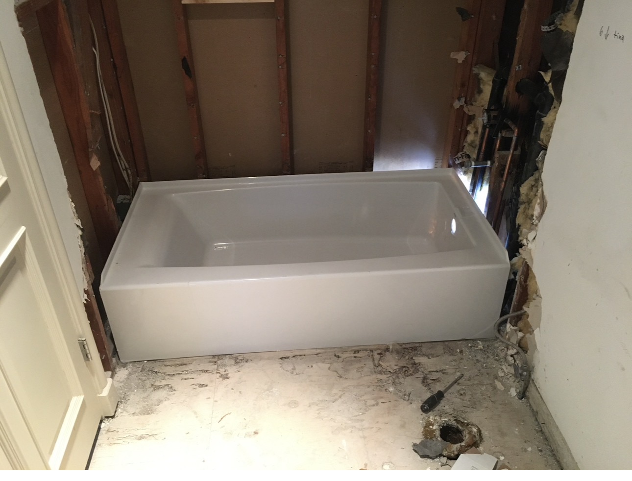 New white bathtub set in place with open stud walls and exposed supply and drain plumbing during a bathroom remodel
