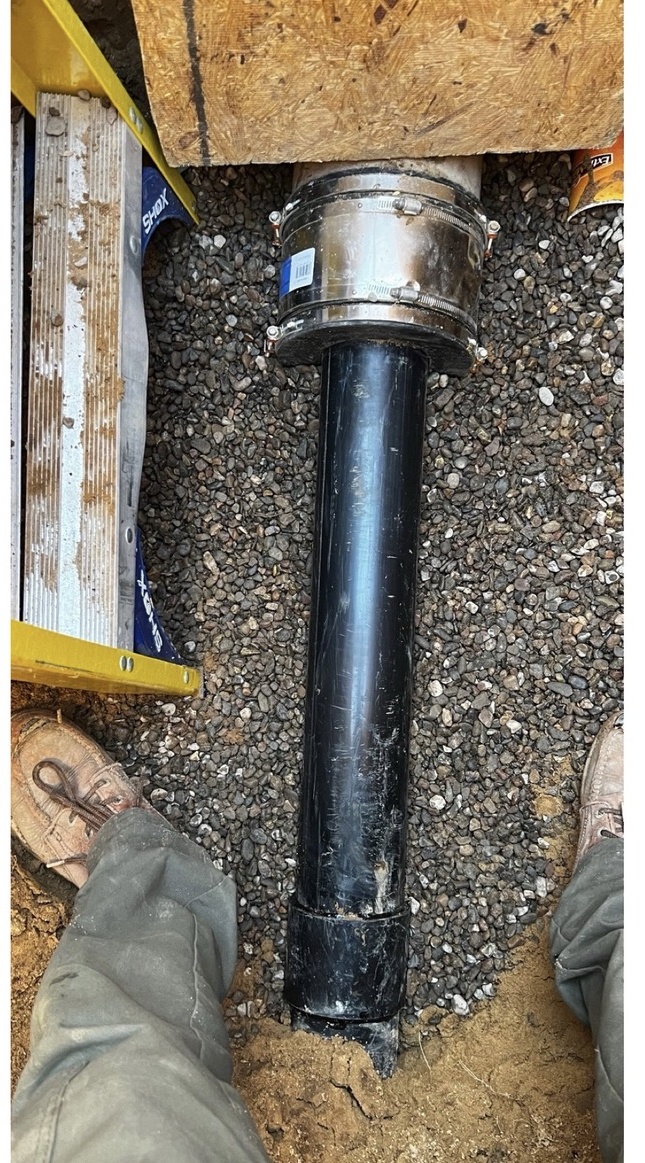 Black sewer pipe joined with stainless banded couplings set in gravel below a floor framing