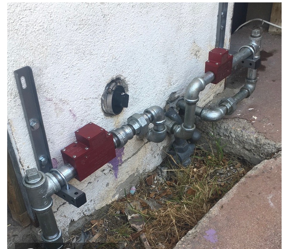 Two red earthquake safety valves on exterior galvanized gas piping running along a building wall