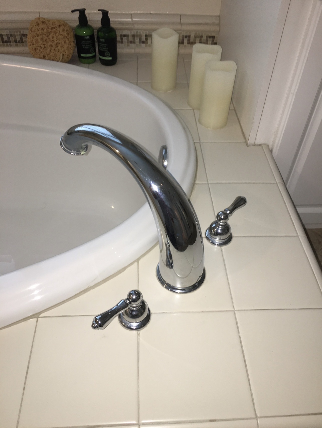 Chrome deck-mounted tub faucet with two lever handles on a white tiled tub surround