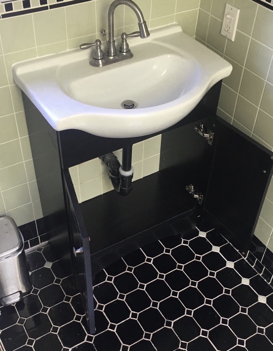White sink on a black vanity with a brushed nickel faucet and exposed drain piping in a tiled bathroom